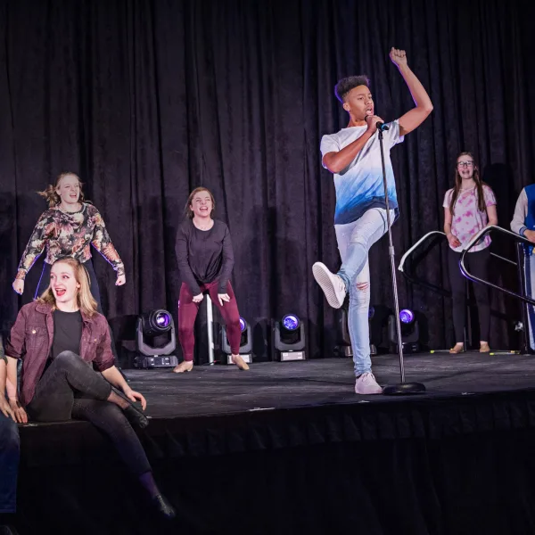 Several actors sitting and standing onstage while singing, with one actor grabbing a microphone on a stand