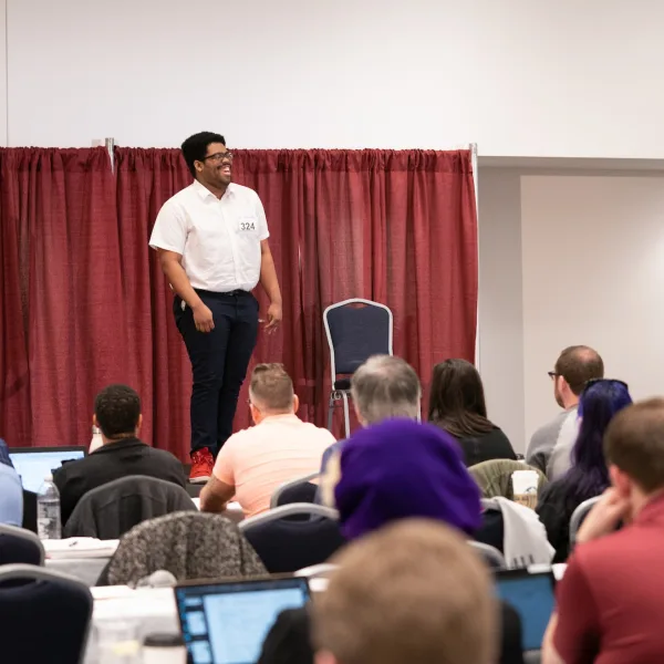 An actor smiling and laughing while standing on a stage in front of auditioners who are seated at tables.